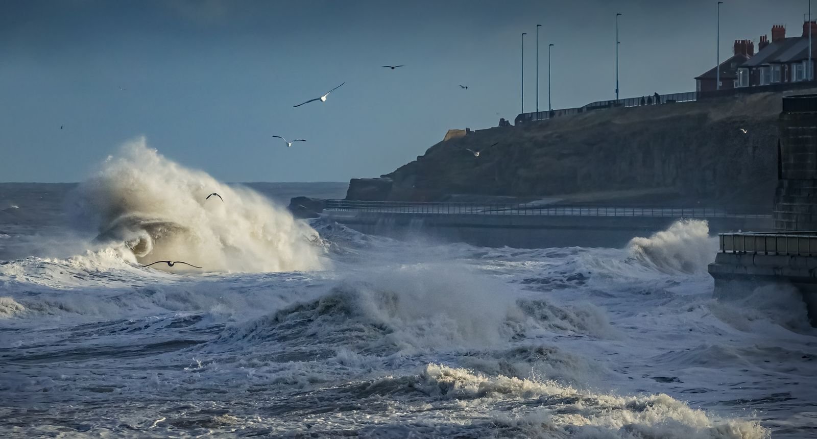 Sturmwellen brechen an einer Uferpromenade an der Nordsee — Möwen im Wind über der Gischt